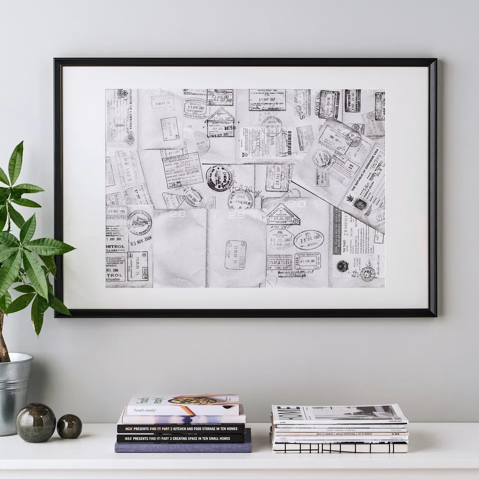 Framed travel-themed black and white art with stamps, mounted on a wall. Surrounded by books and a plant on a shelf.