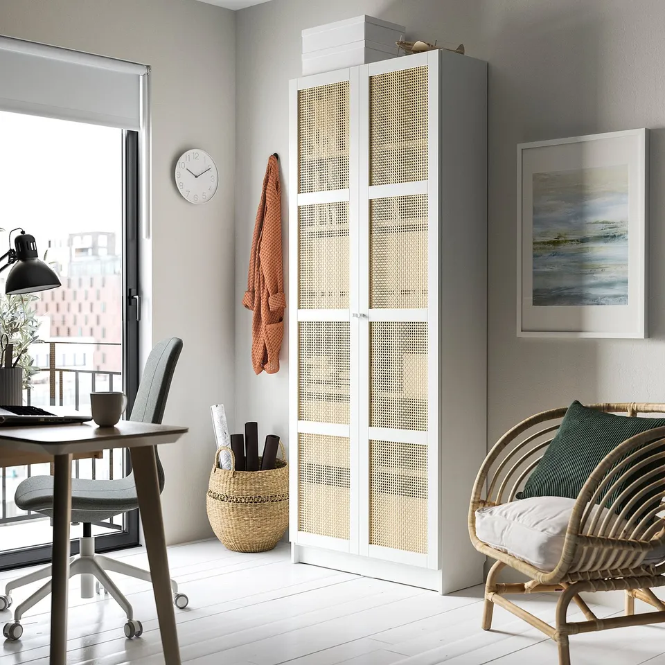 Modern home office with white BILLY bookcase, desk, chair, and clock. Woven bamboo doors, cosy wicker chair with pillow.