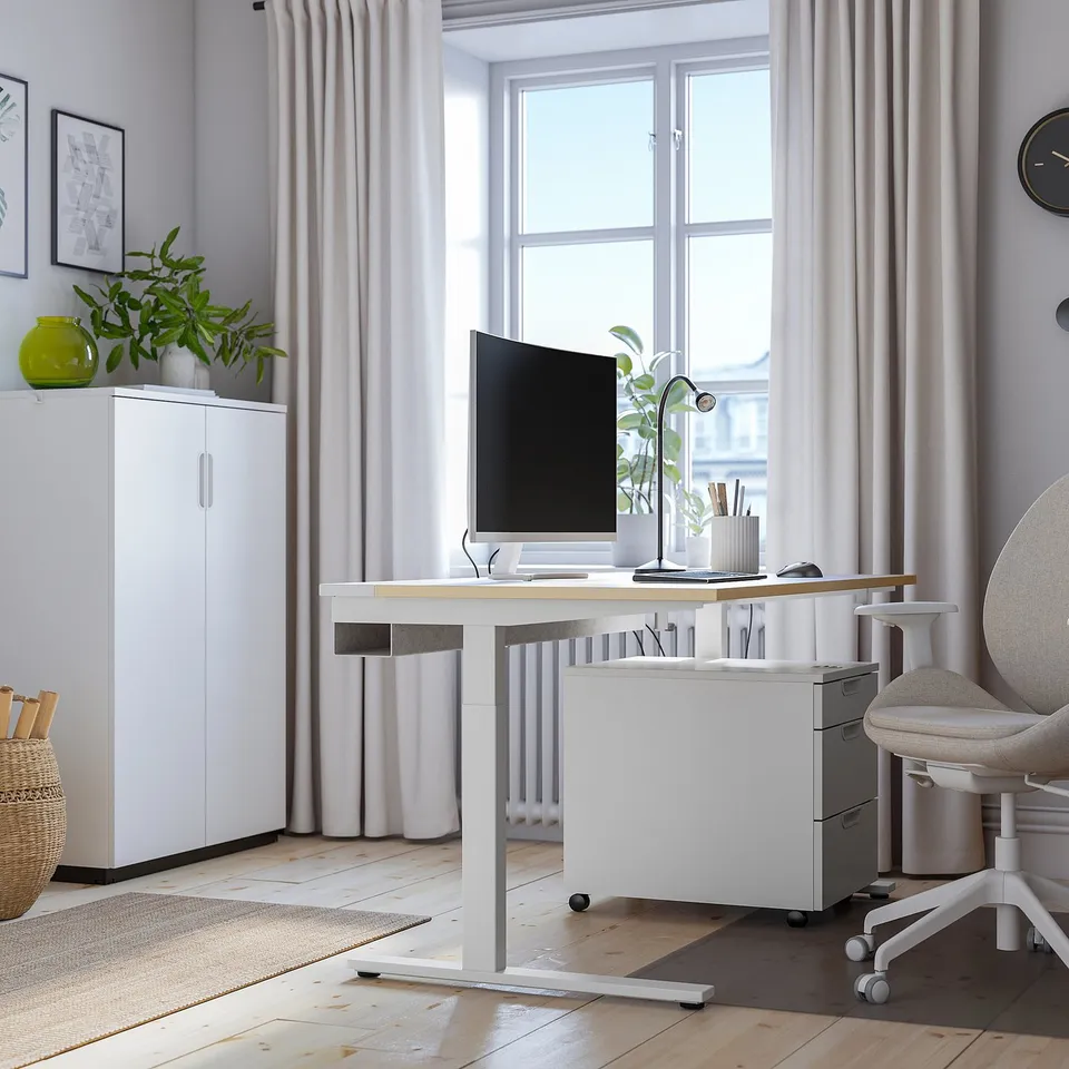 Modern home office with height-adjustable desk, computer, grey chair, and window. White storage, basket, light wood top, bright and clean.