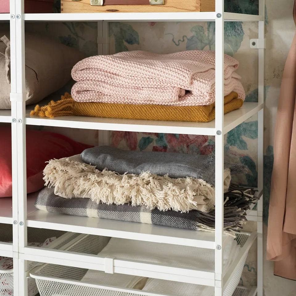 White JONAXEL storage shelves holding pink, grey, and yellow folded blankets. Organised, multi-use design.