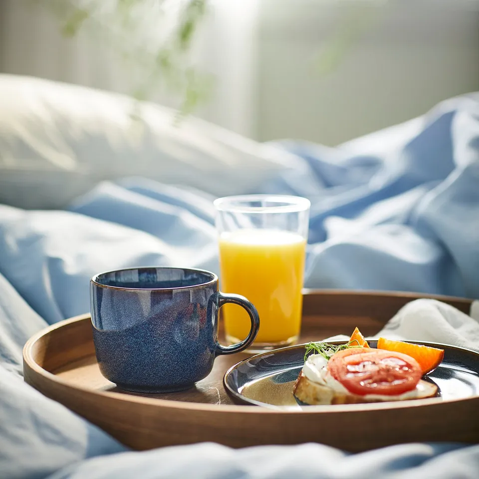 Morning breakfast on bed, featuring a GLADELIG mug in ocean-blue with gold rim, wood tray, juice, & tomato toast.