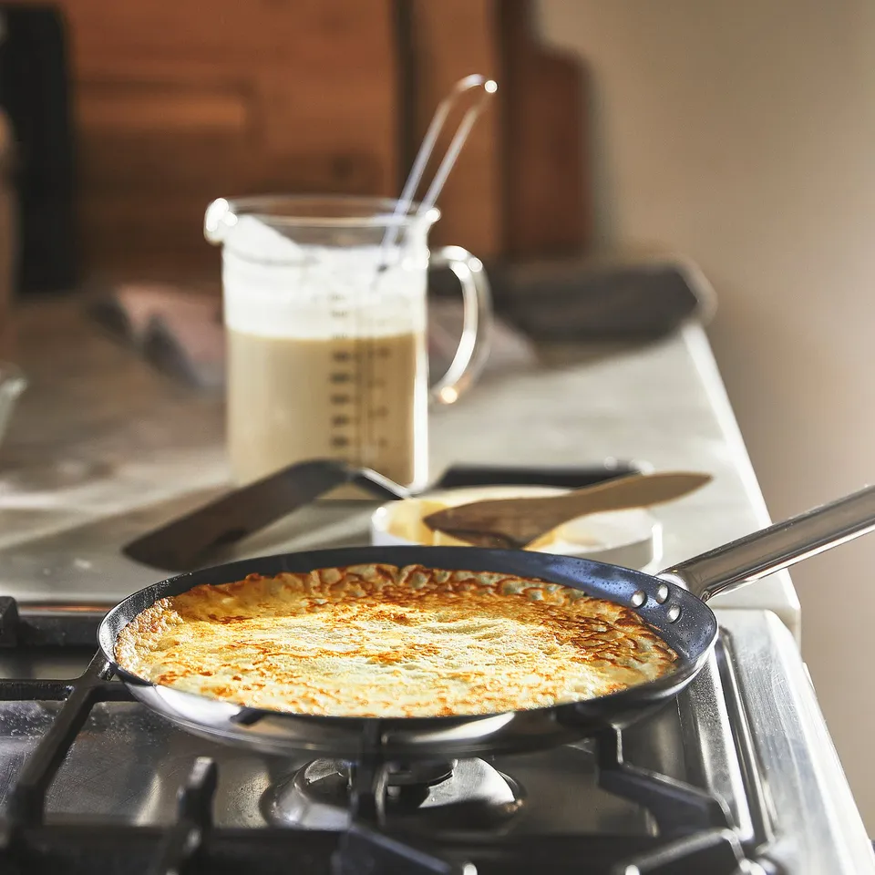 A round frying pan on a stove with a golden-brown crepe inside, next to a jug with batter. The pan is on a heat-resistant surface.