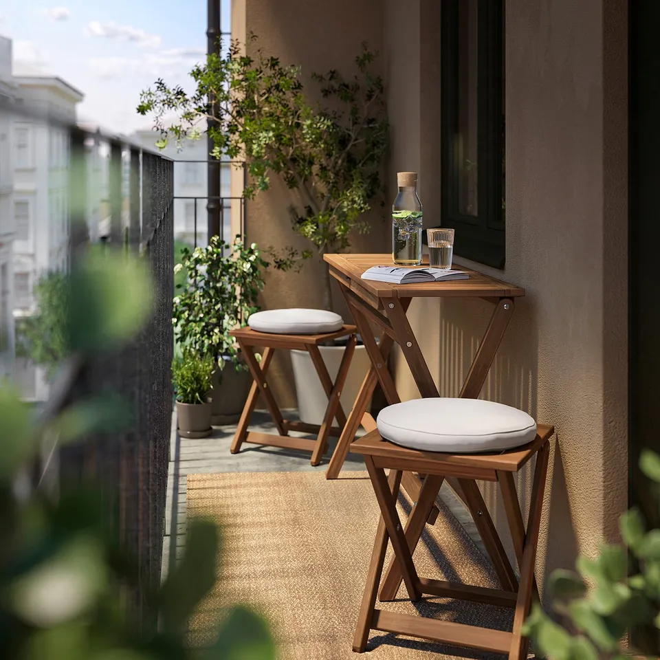 Balcony setup with NÄMMARÖ brown acacia wood table and stools, white cushions, glassware, and greenery.