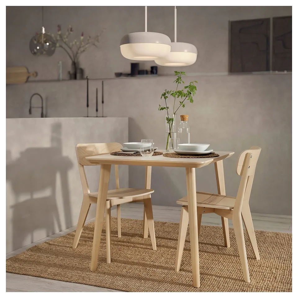 Modern light wooden dining set with table, chairs, plates, glasses, and vase with plant.