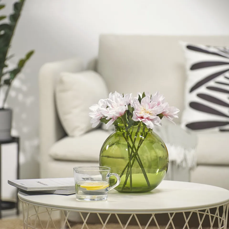 A small green recycled glass vase with pink flowers sits on a white coffee table, accompanied by a tea cup and magazines.