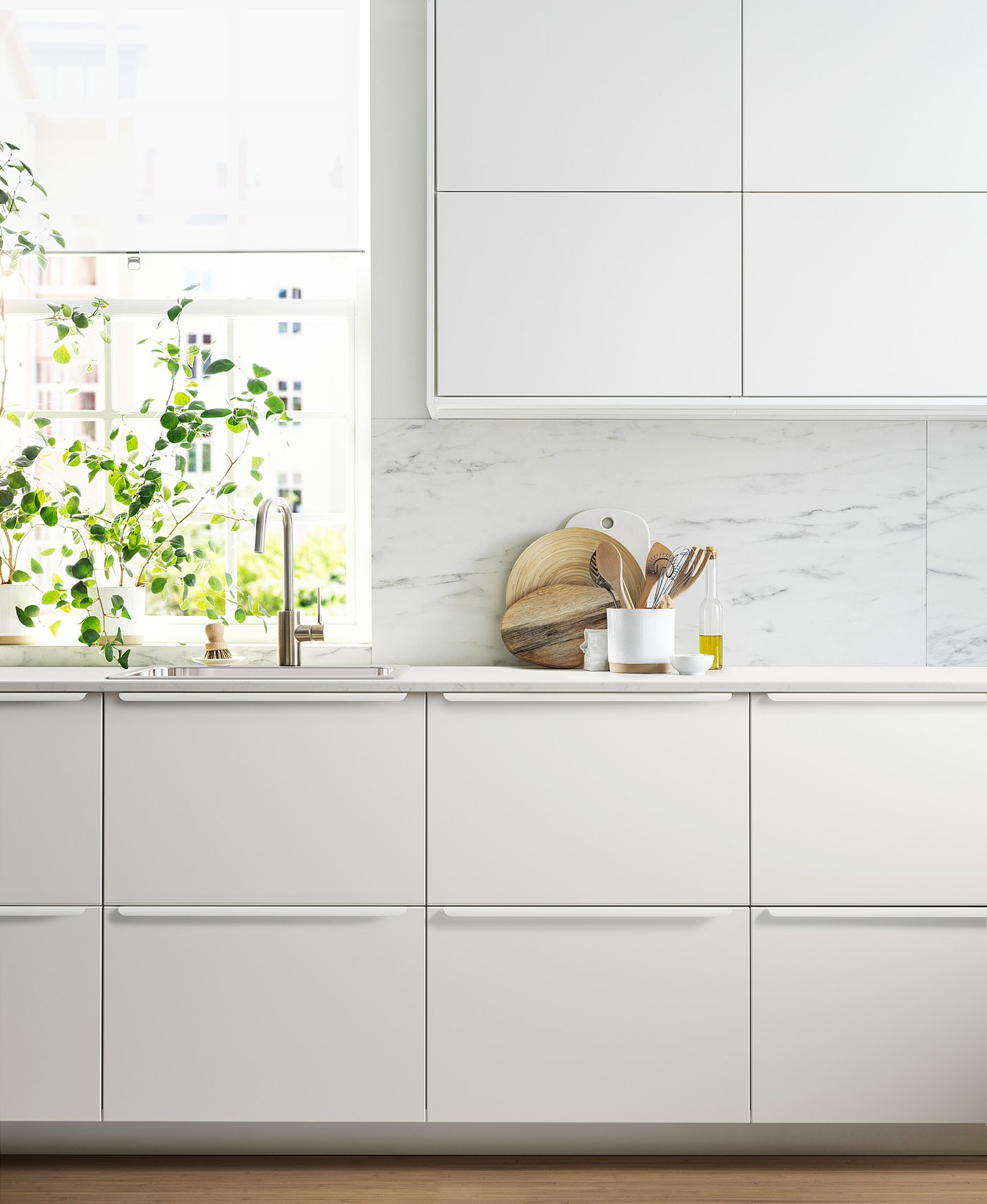 Modern white kitchen with marble backsplash, sleek cabinets, stainless sink, and green plant by window.