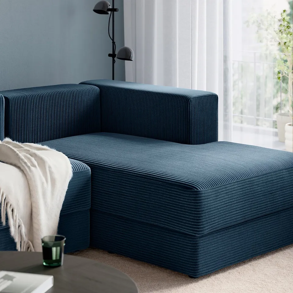 A modern, modular sofa sits by a window. Its chaise longue offers relaxation, with a cosy throw blanket draped over its side.