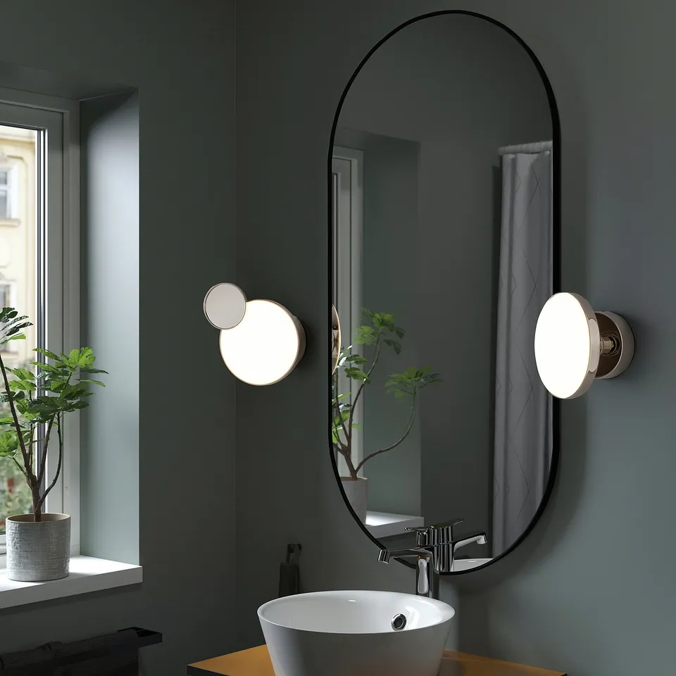 A modern bathroom with a KABOMBA wall lamp featuring a removable mirror, sleek black frame, circular lights, and a small plant on the window sill.