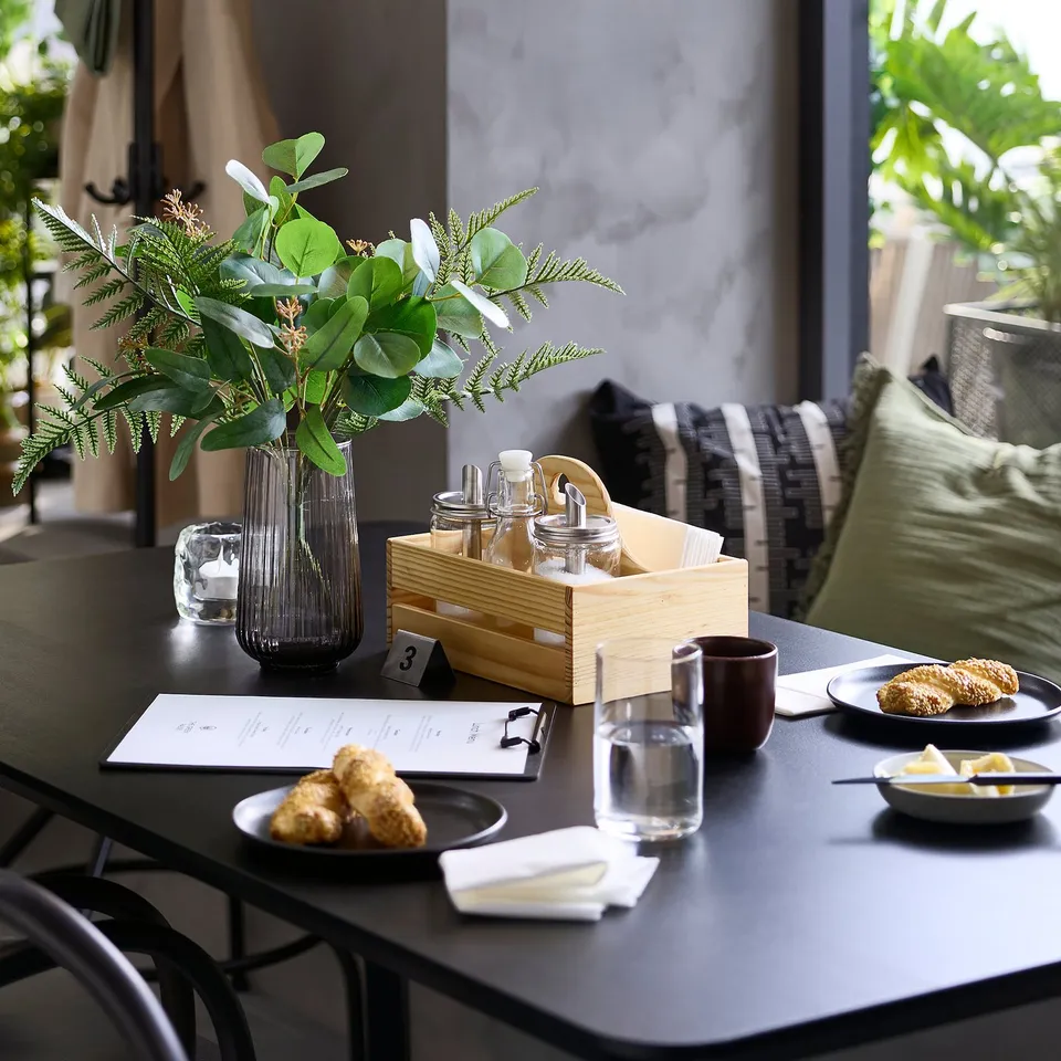 Black table with spice jars, greenery, food; wooden box with bottles; clear vase with plants; plates and cups.