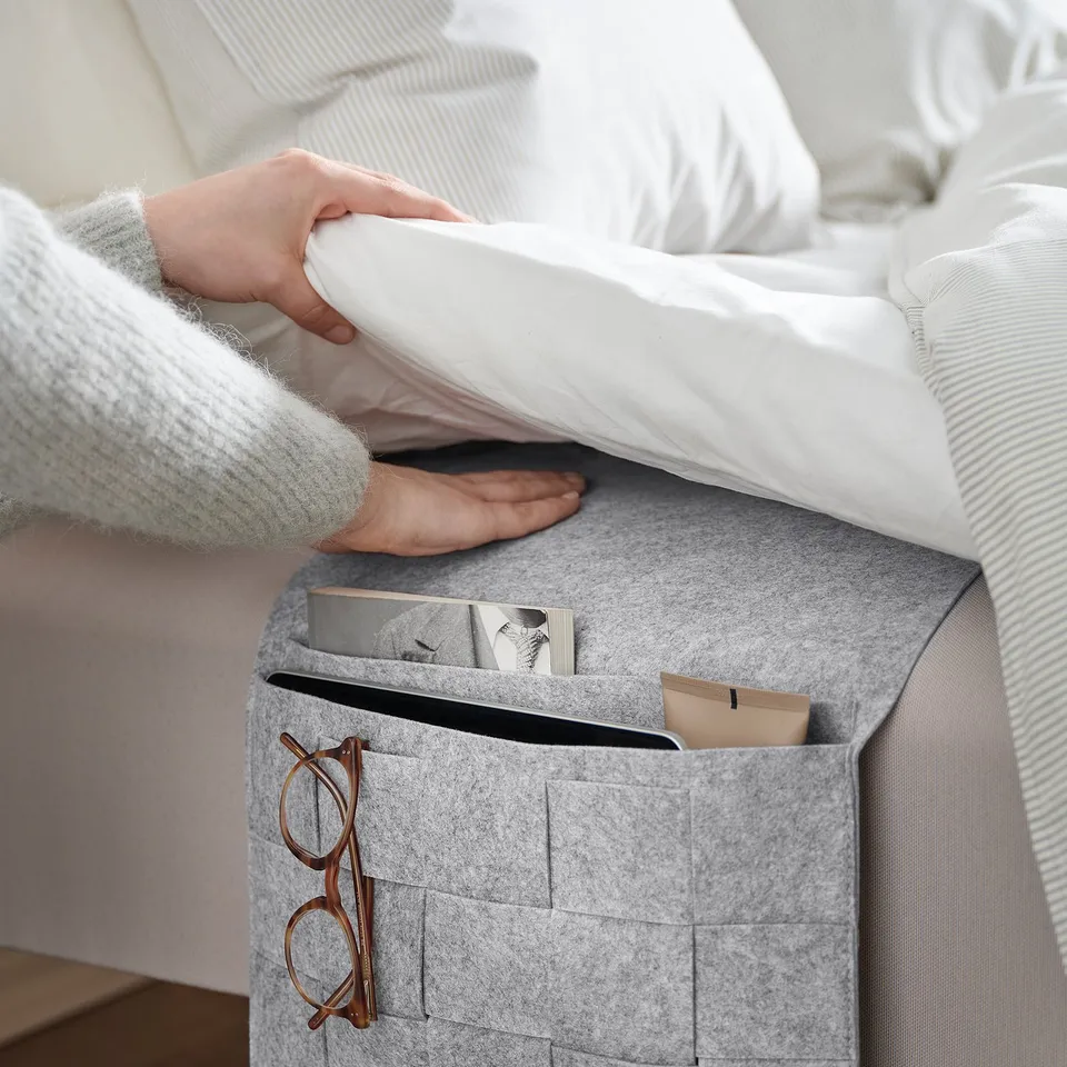 A person in a grey sweater sits on a bed beside a grey felt storage pocket with a chequered pattern and mesh squares, holding a white pillow.
