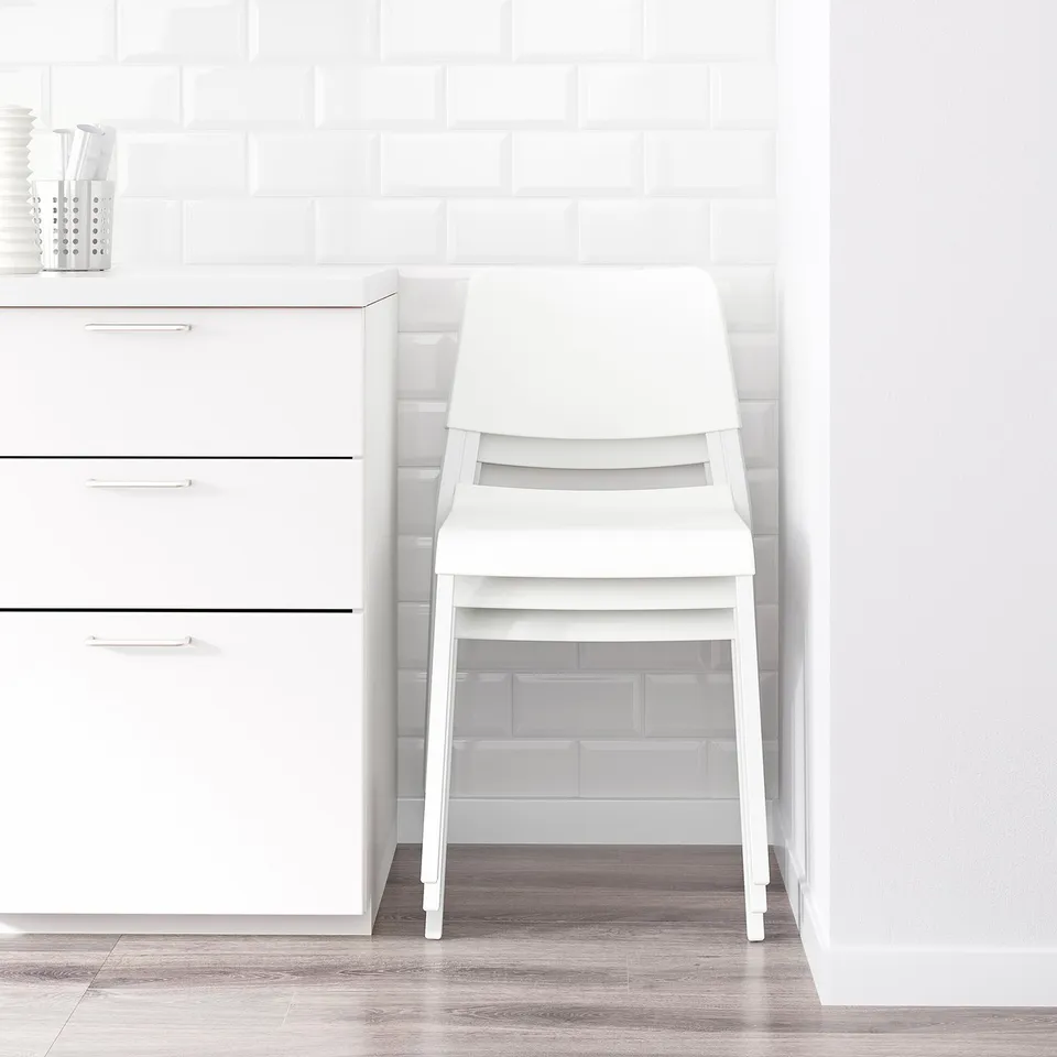 Clean, minimalist kitchen scene with white NORDEN table and stacked chairs neatly stored against white cabinet. Modern, space-saving design.