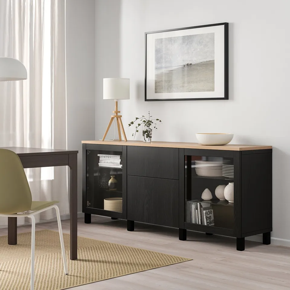 Black ikea BESTÅ sideboard with glass doors, wooden top, next to lamp & plant.