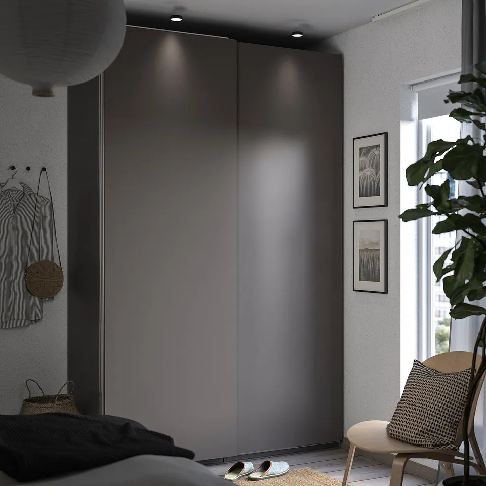 Modern bedroom with dark HASVIK wardrobe, white walls, plant, chair, and sandals.
