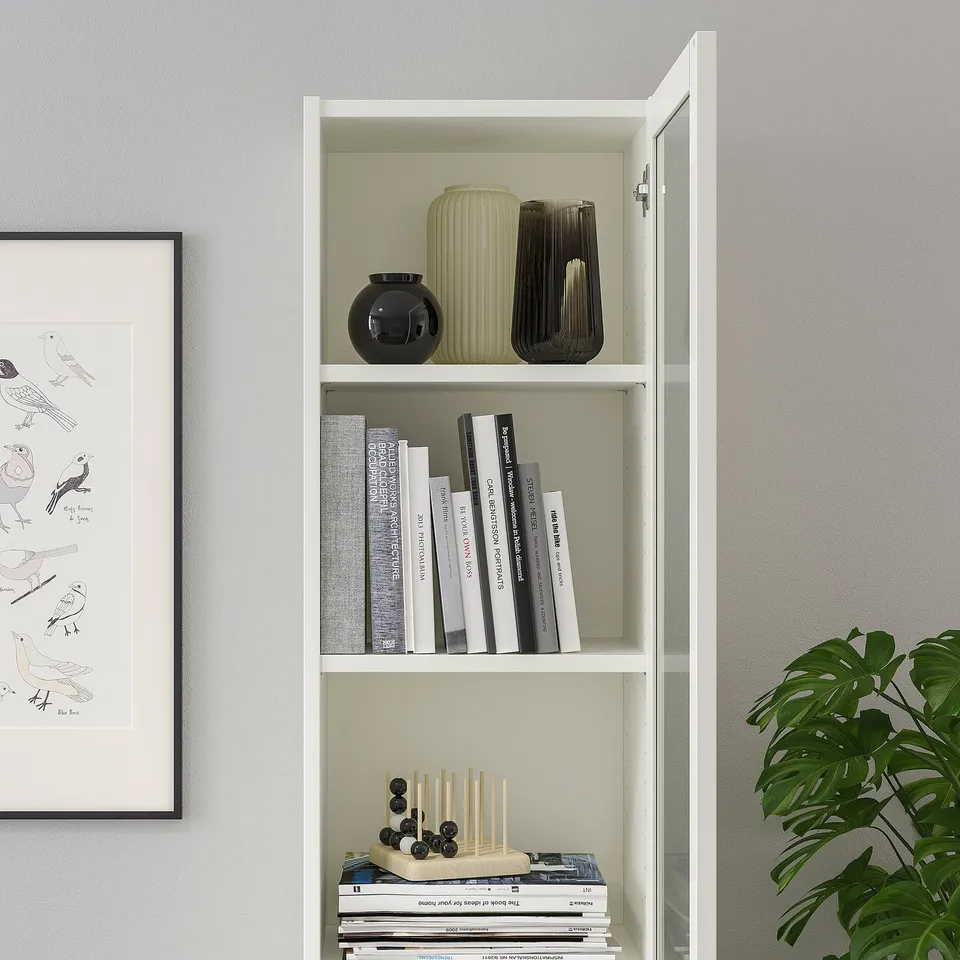 White BILLY bookcase with glass door, displaying books, vases, and decorative items, mounted on wall.