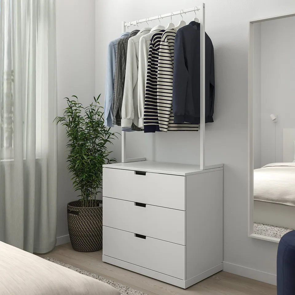 A NORDLI white chest of drawers with a clothes rail on top, placed next to a window with a plant and a bed.
