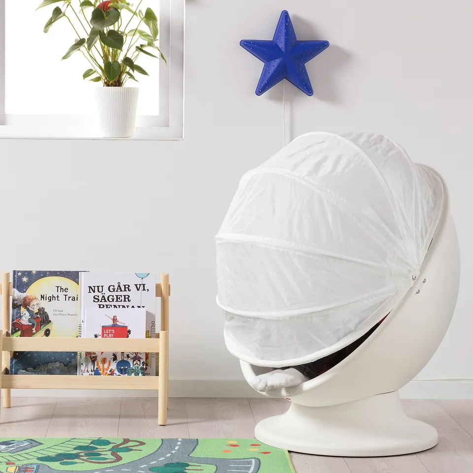 Cosy white IKEA PS LÖMSK kids armchair with hood, next to bookshelf with colourful books, blue star light, and green plant.