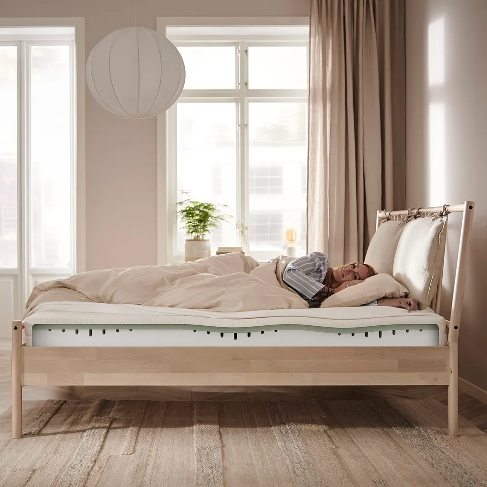Wooden MALM bed with person sleeping. Beige linens, high headboard, near windows with curtains and pendant light.