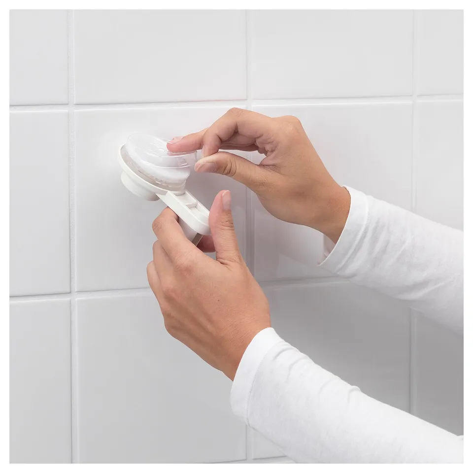 A person attaches a clear, white-framed soap dish with suction cup to a tiled wall. The dish is removable for easy cleaning.