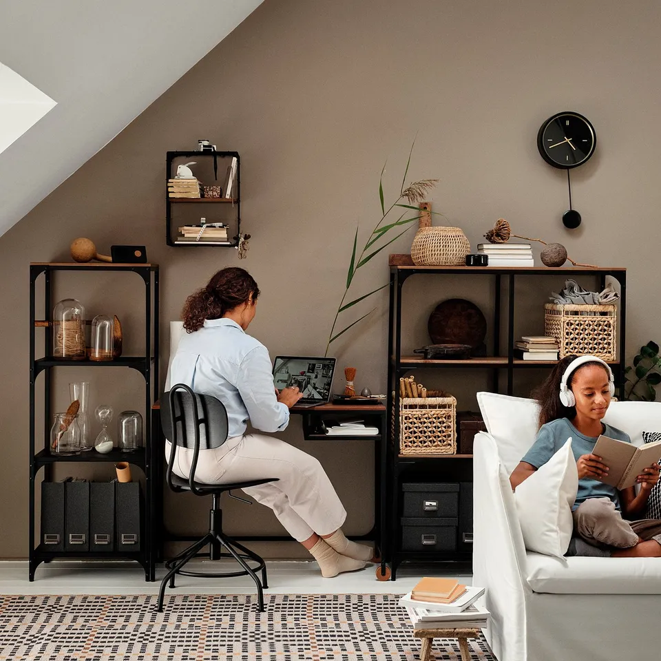 Two people in home office: one at desk, other on couch with headphones. Modern decor with books and vases.