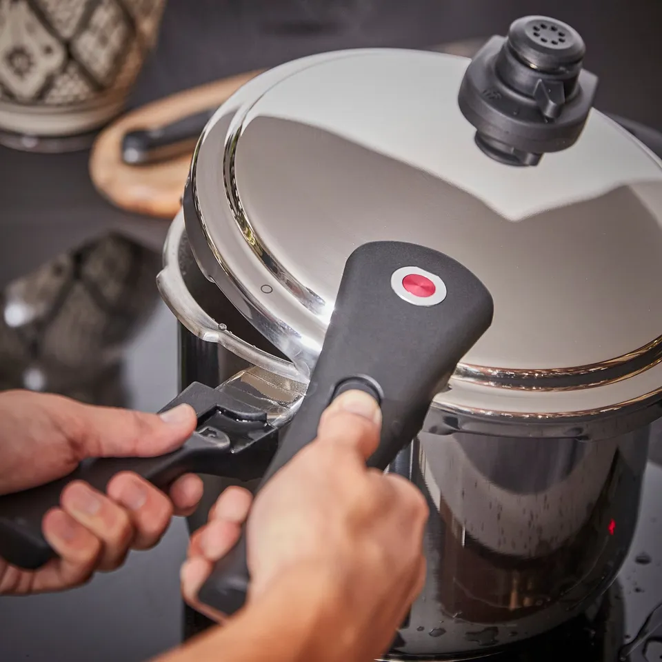 A person operates an IKEA 365+ stainless steel pressure cooker in a kitchen. The cooker has black handles and a red indicator button.