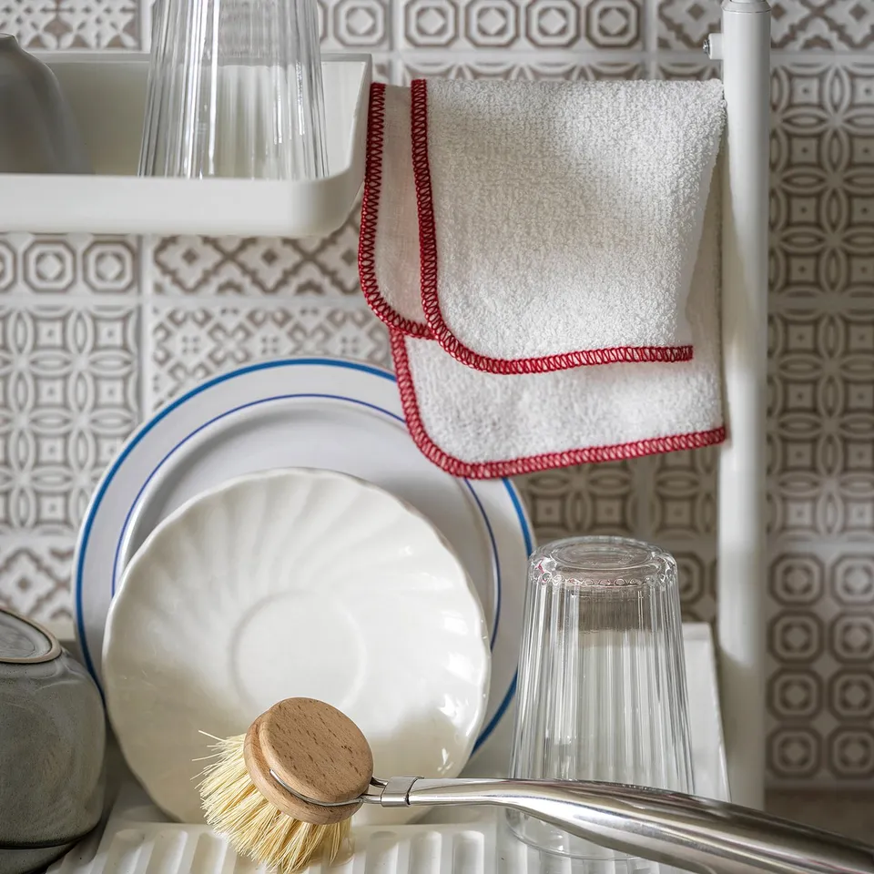 White SUNNERSTA kitchen rack with dishes, glass, and brush. Grey tiled wall, red-edged towels.
