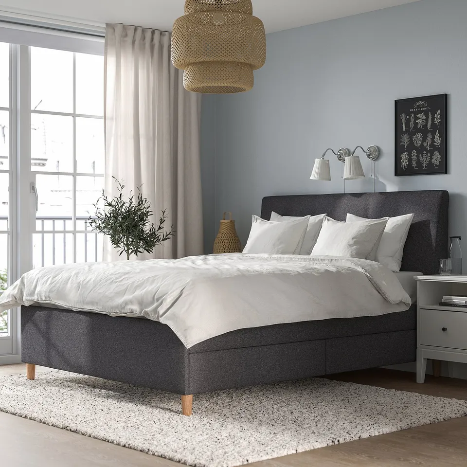 Modern bedroom with ÅRSTID lamp overhead. Grey bed, white bedding, plush rug. Light blue walls, natural light from window.