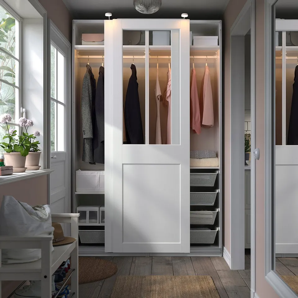 White PAX wardrobe with glass doors displaying clothes and storage. Decorative flowers on nearby windowsill.