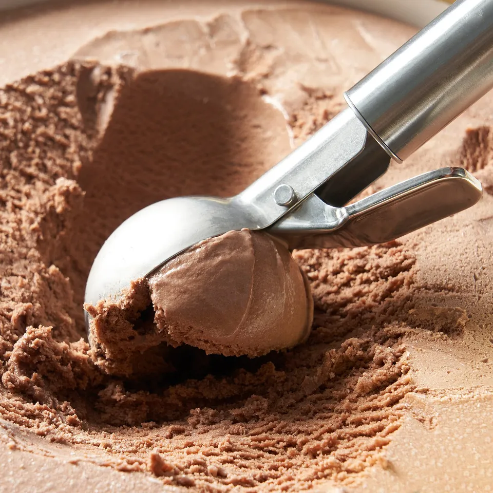 Metal ice cream scoop holding chocolate ice cream, pressing to release a perfect round scoop into a container.