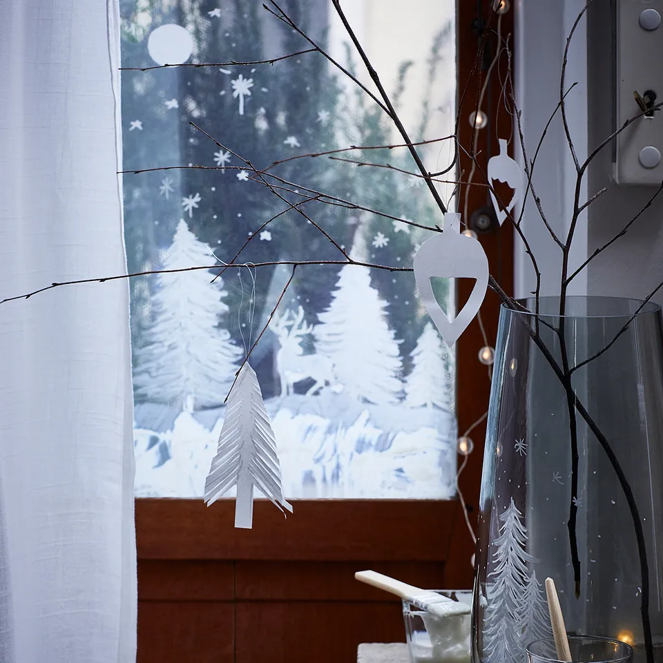 Windowsill decorated with white paper christmas trees, a jar of branches, and fairy lights. Wooden cabinet, white curtain, and drawing tools nearby.