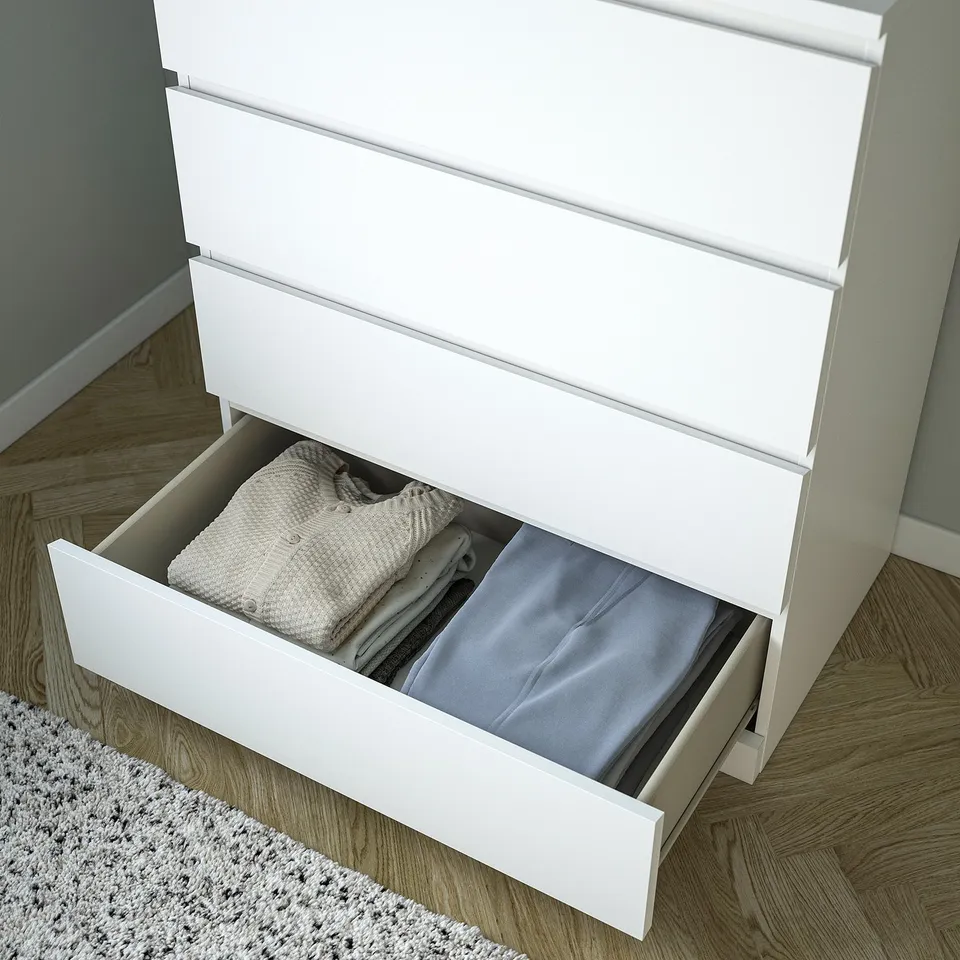 Open white MALM dresser with folded clothes inside. Drawers are smooth and can store multiple items. Safety feature noted as wall-anchoring.