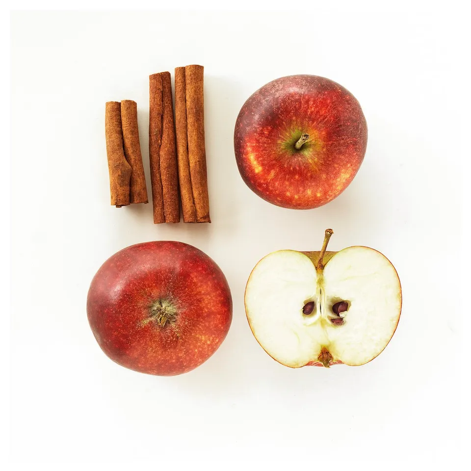 Apple-shaped candle with cinnamon sticks, whole apple, and halved apple image. Red apples, white background, arranged in a square.