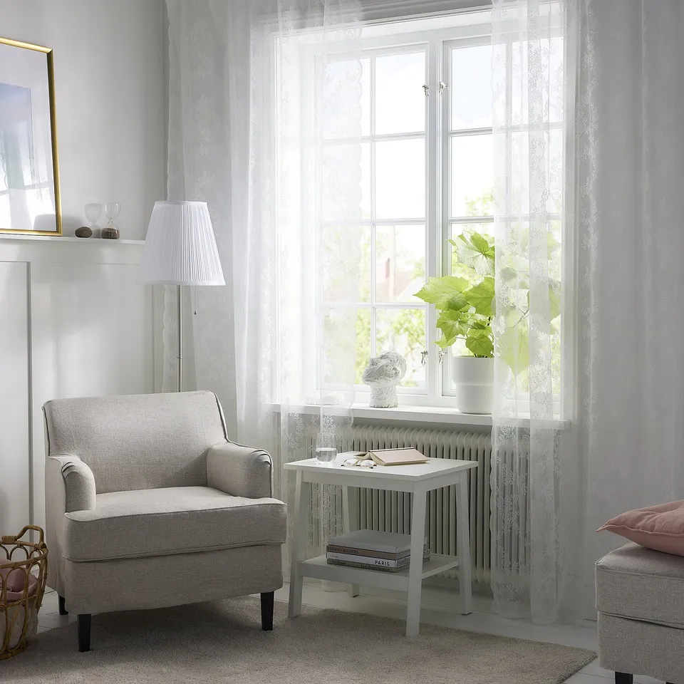 Bright living room with lace curtains, armchair, and table. Natural light and greenery visible through curtains, creating cosy modern space.