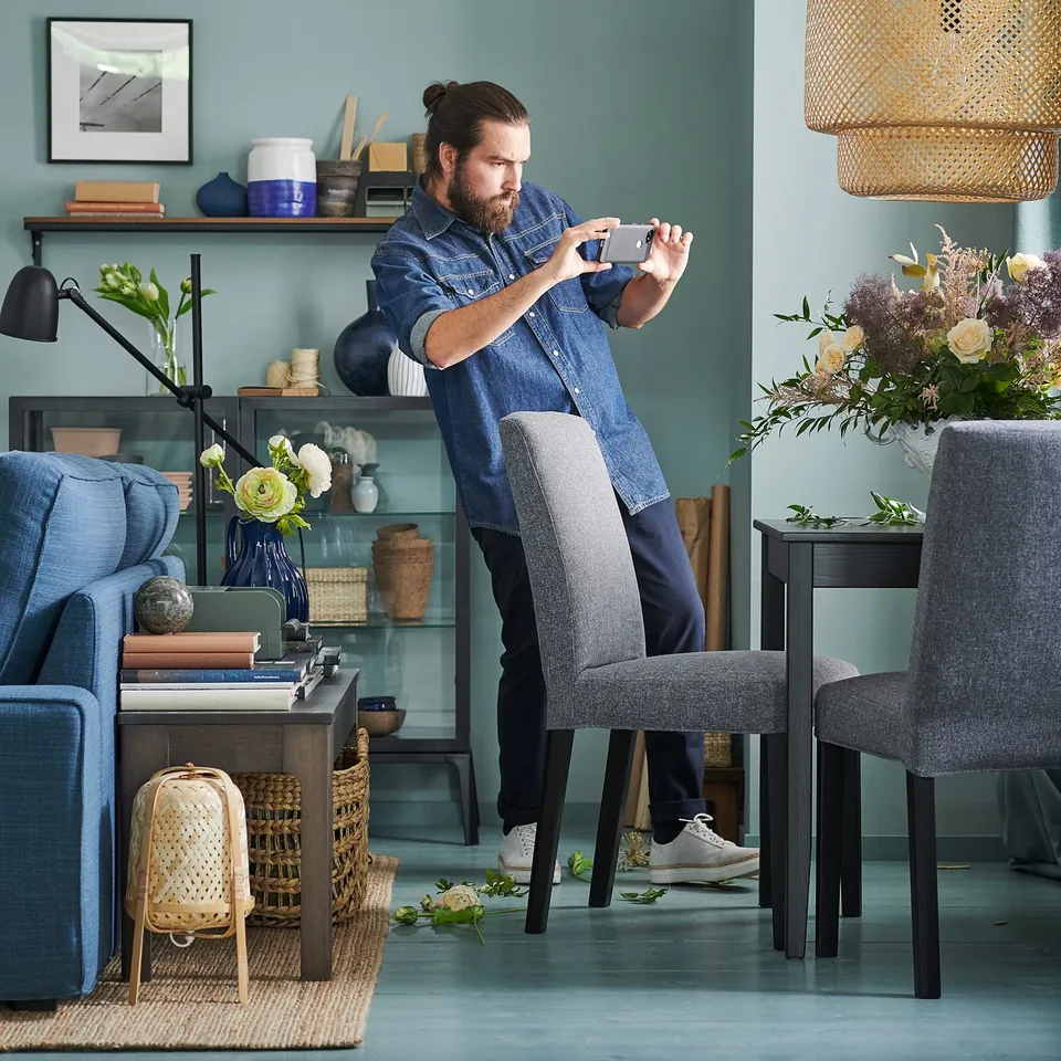 Person taking photo in stylish room with blue walls, a blue BERGMUND chair, and wooden dining table with grey chairs.