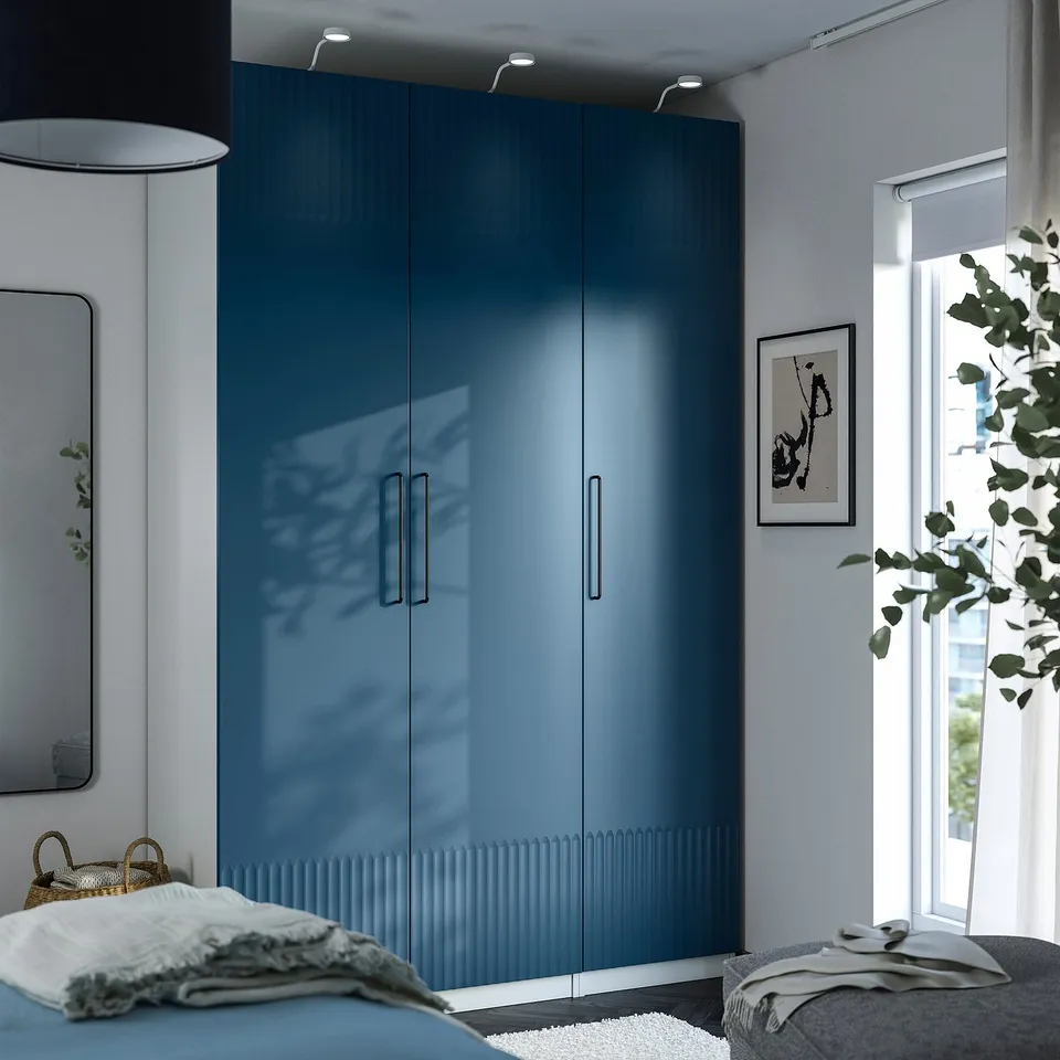 Blue PAX wardrobe by window with lamp. Bed, mirror, plant in minimalist room.