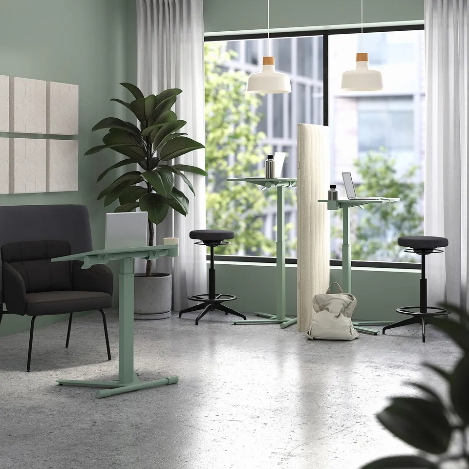 Modern office space with mitzon green height-adjustable laptop tables, black stools, grey lounge chair, large plant, and big window.