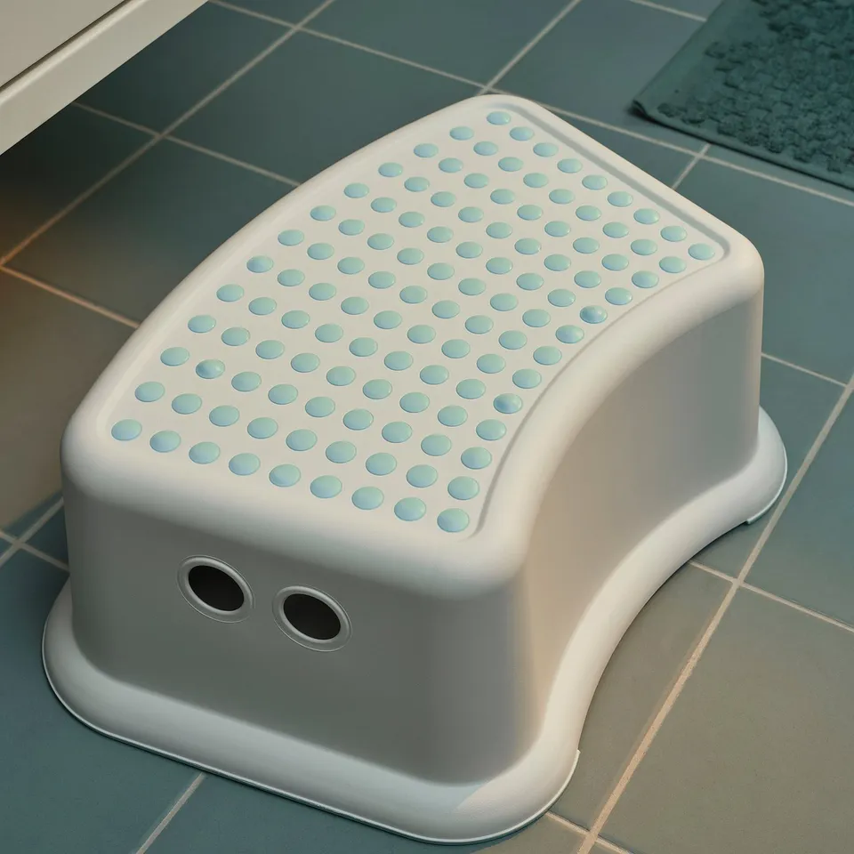 A white FÖRSIKTIG step stool on blue tiles. It has a curved design, anti-slip top with dots, two holes on the side.