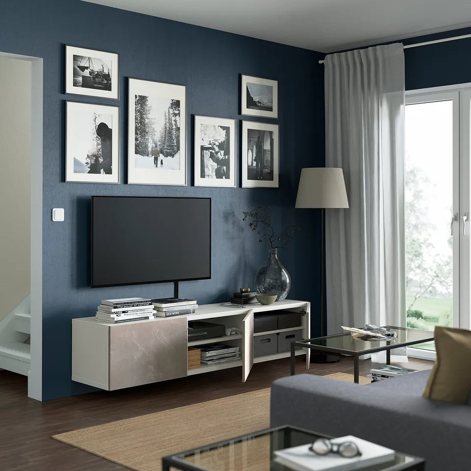 Modern living room with dark blue walls, flat-screen tv, marble stand, books, vase, glass coffee table, natural light, and black & white photos.