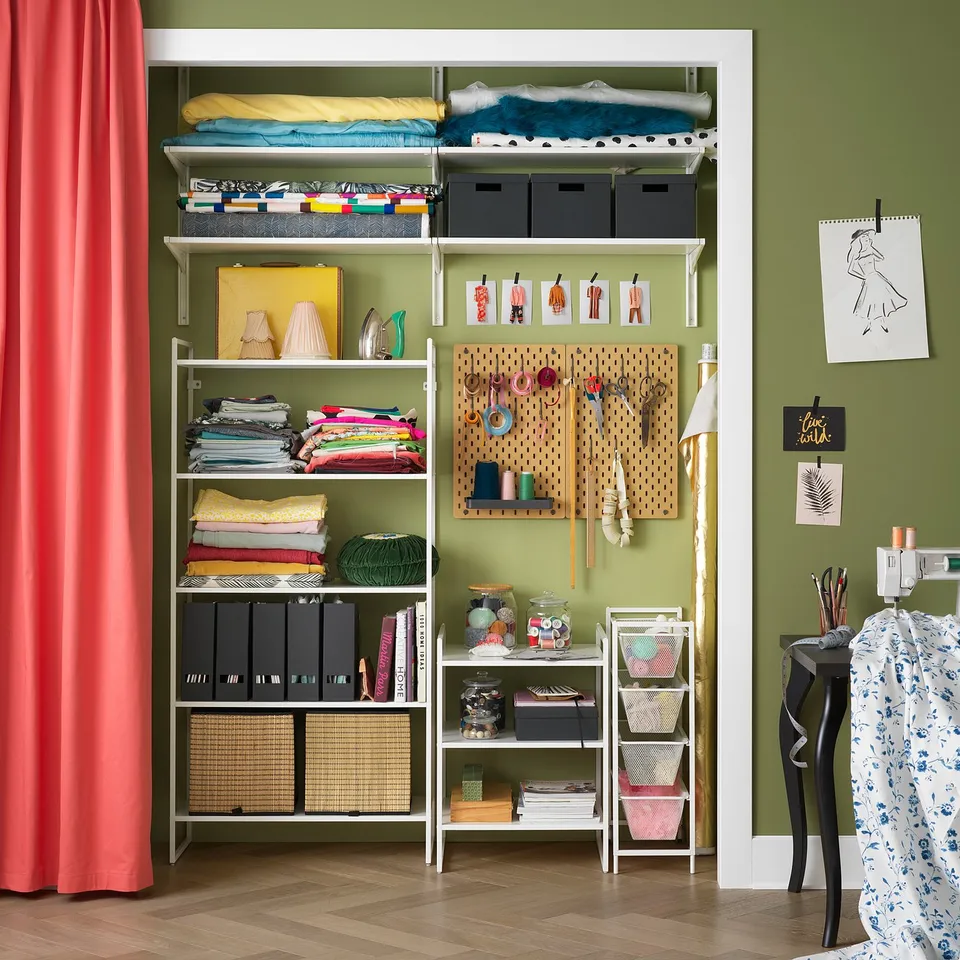 Organised craft room with white JONAXEL storage shelves holding fabrics, books, and supplies against green wall.