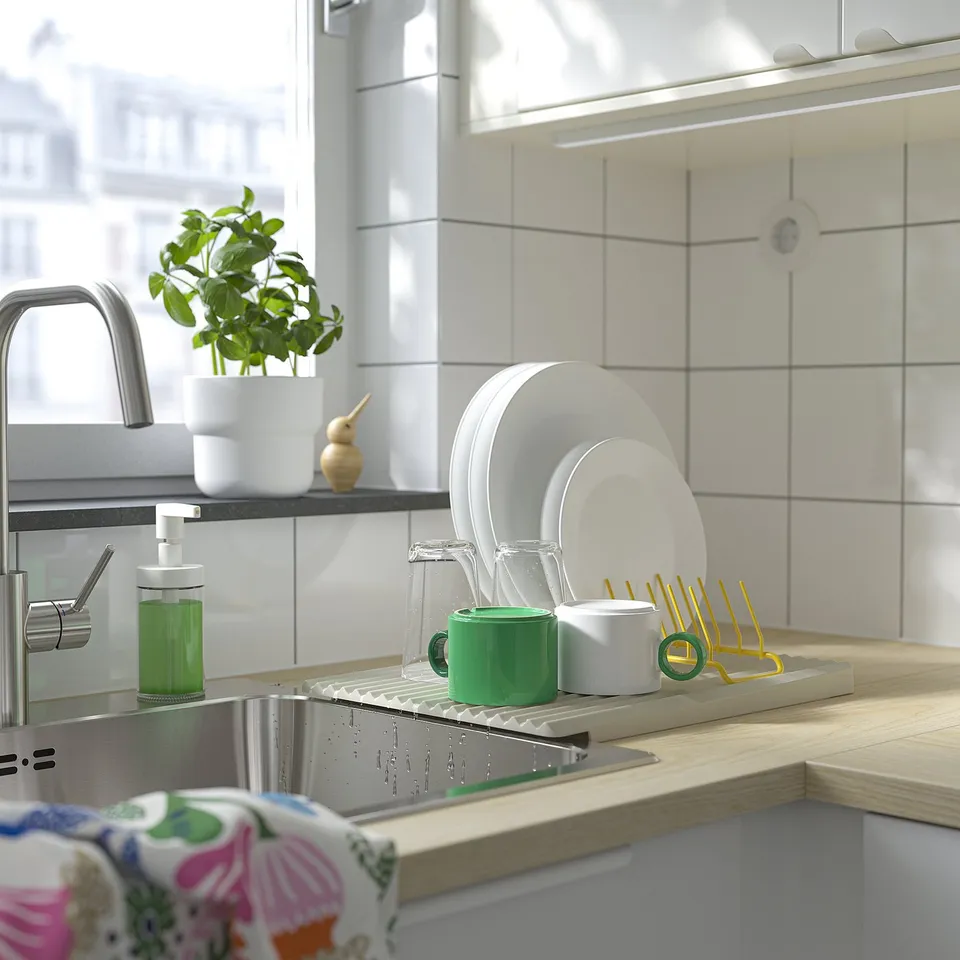 A kitchen sink with plates in a yellow drying rack. A green cup and soap dispenser are visible. Water droplets are present, indicating recent use.