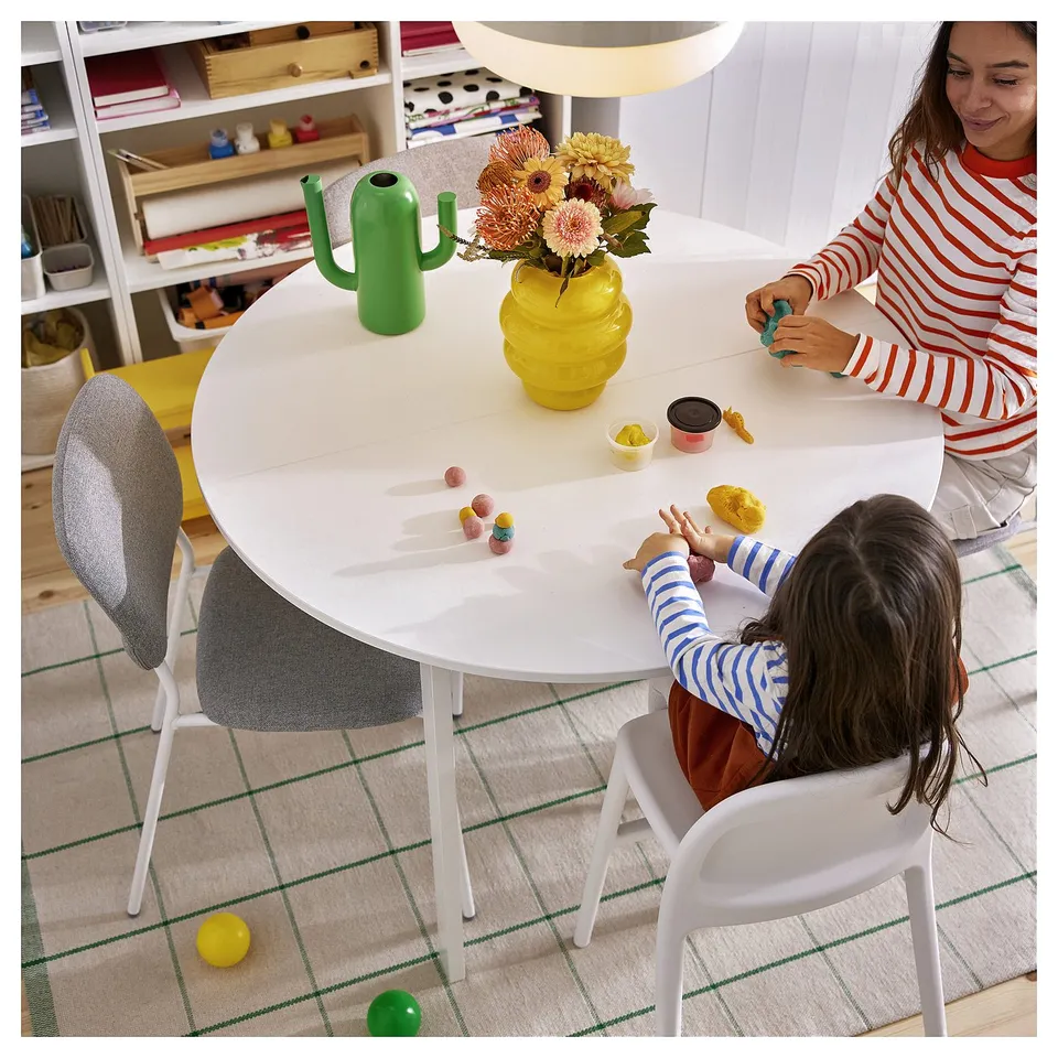 Two people at table with green/white rug, yellow vase, cactus bottle, and bookshelf. Person in stripes, child in blue/white.