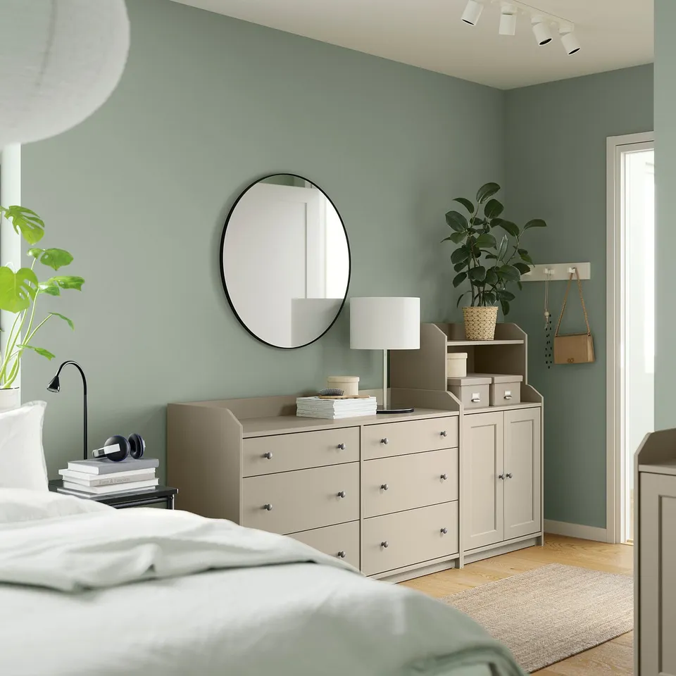 Bedroom with green walls HAUGA dresser and drawer combo in beige, topped with a lamp and plants. Round mirror above, white bed linens below.