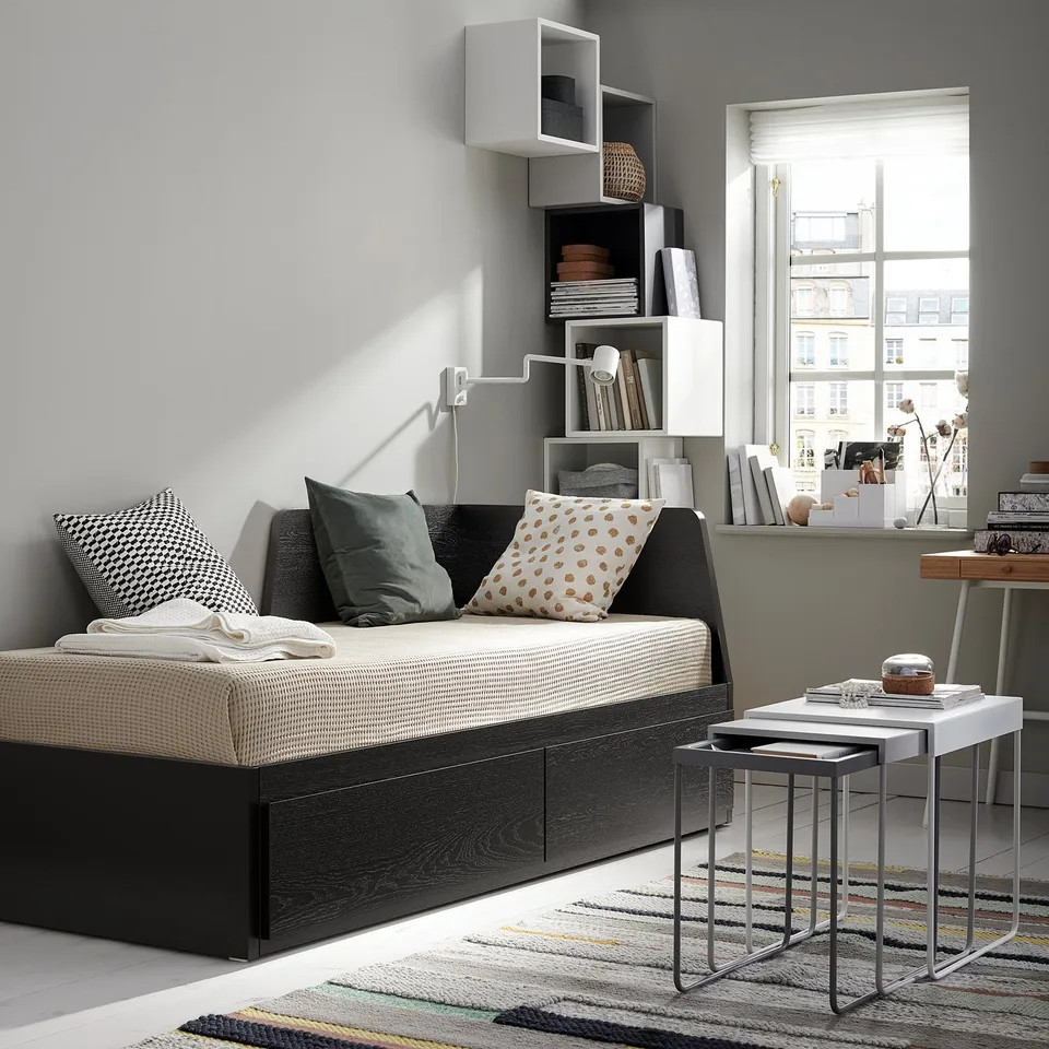 Modern living room with a FLEKKE day-bed, white shelves, colourful rug, and metal nesting tables.