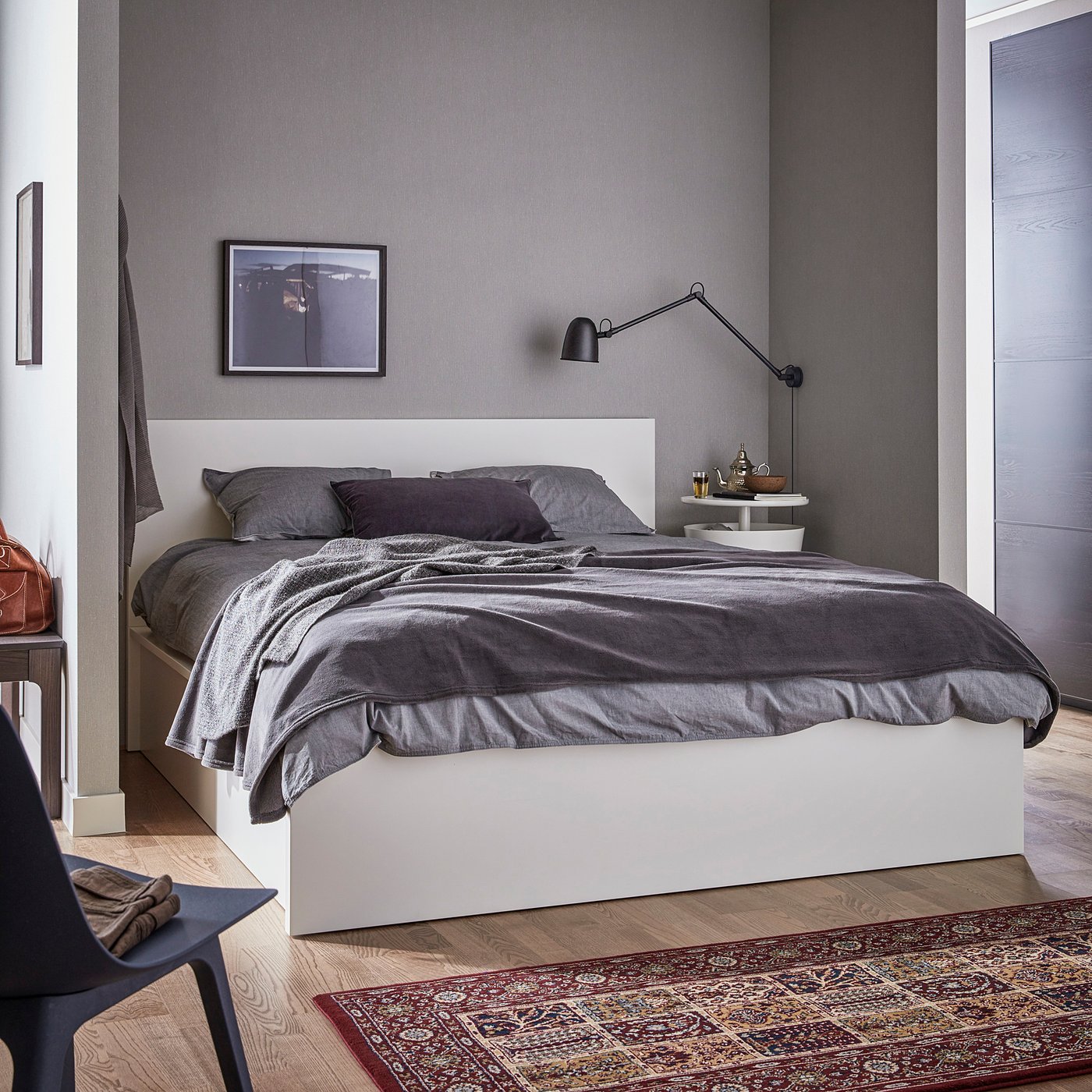 Modern minimalist bedroom a white MALM bed with grey linens, grey walls, and a black moveable lamp. A colourful rug lies on wooden floors.
