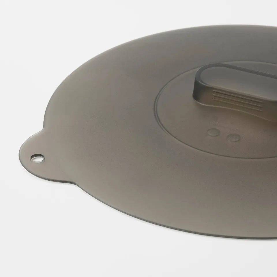 Round, dark silicone kitchen lid with small handle, lying on a white surface. Durable, multi-use, fits various sizes of pots and pans.