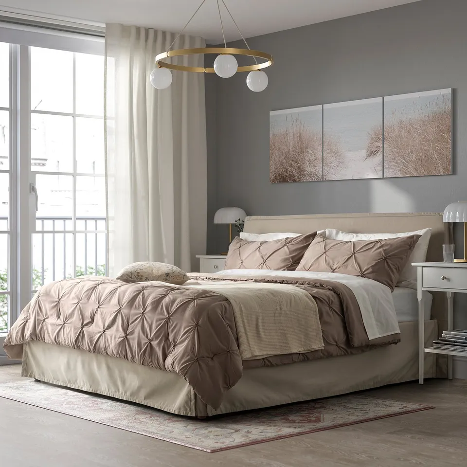 Cosy bedroom with beige BÄGARKLOCKA bedding, gold chandelier, grey walls & rug, beach prints, & bedside table lamps.
