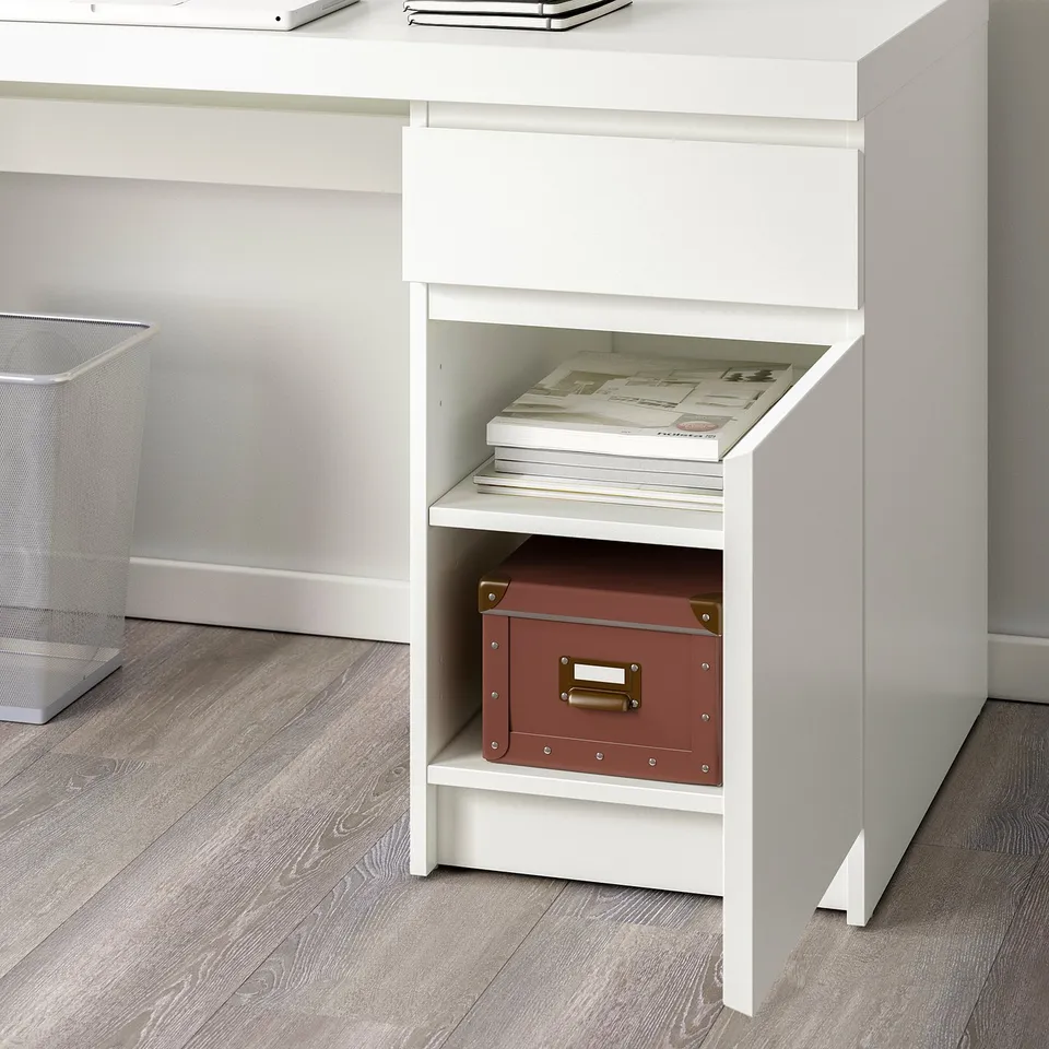 A white desk from the MALM series with a storage shelf underneath holding a red box and stacks of paper.