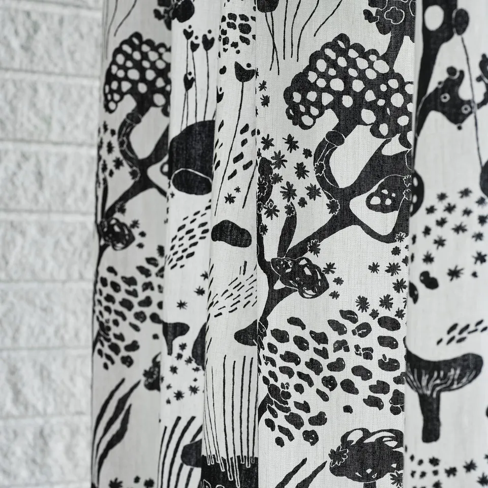 Black & white curtain with nature designs, pleated, against brick wall.