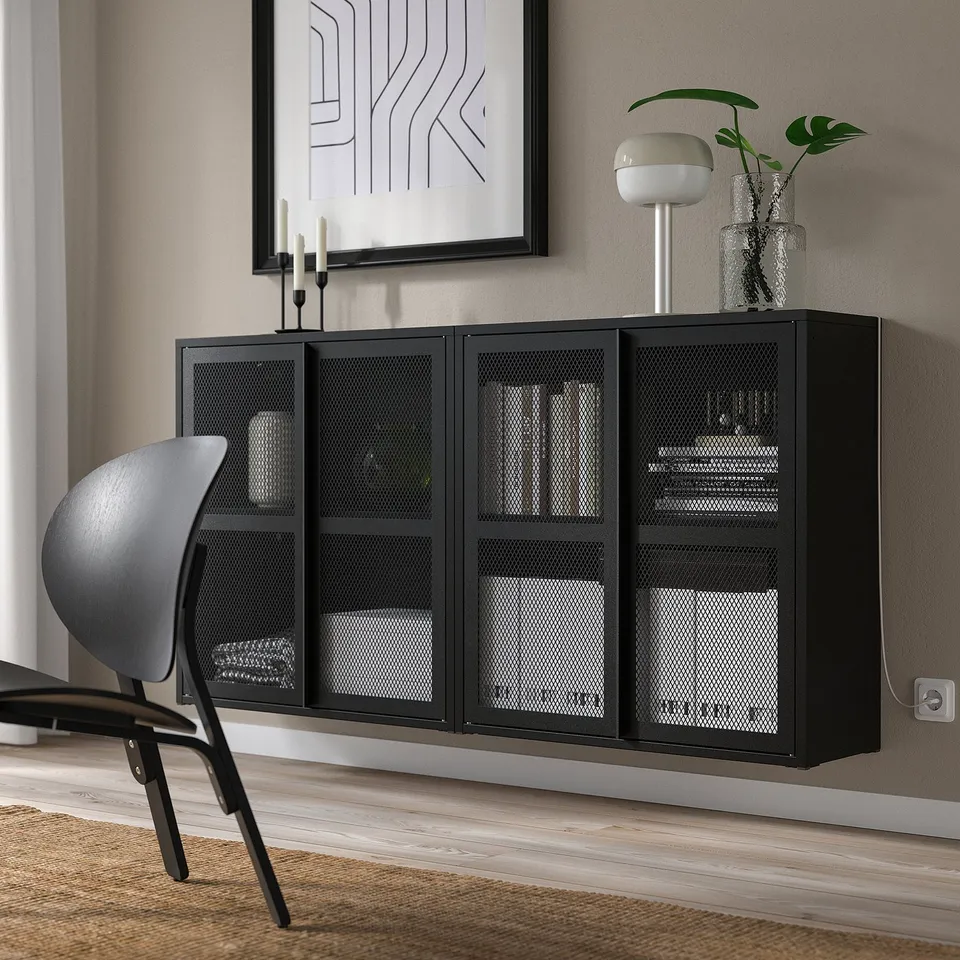 Black metal IVAR cabinet with mesh doors, mounted high on wall, storing books and vases, with grey chair and abstract wall art nearby.