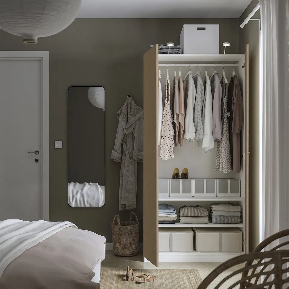 A modern bedroom with a PAX wardrobe open, displaying clothes, shelves with folded clothes, and storage bins. A mirror and a robe hang on the wall.