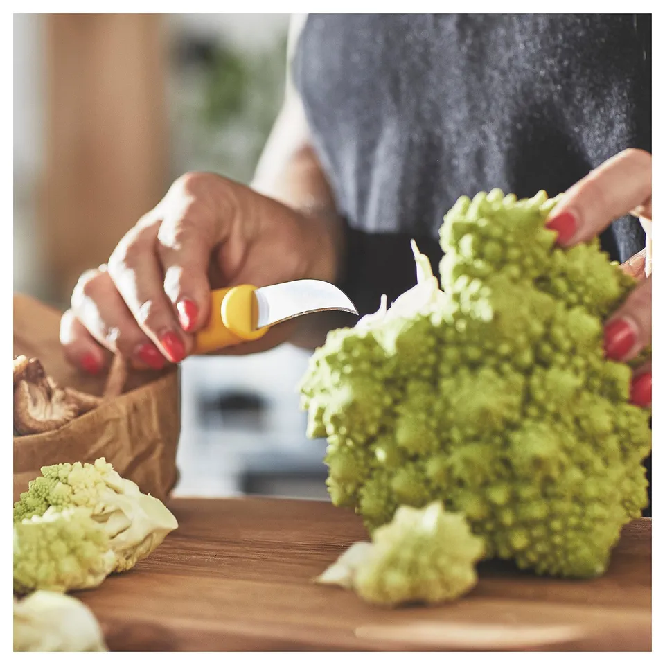 A person peels green veggies with a small, yellow-handled paring knife on a wooden cutting board.